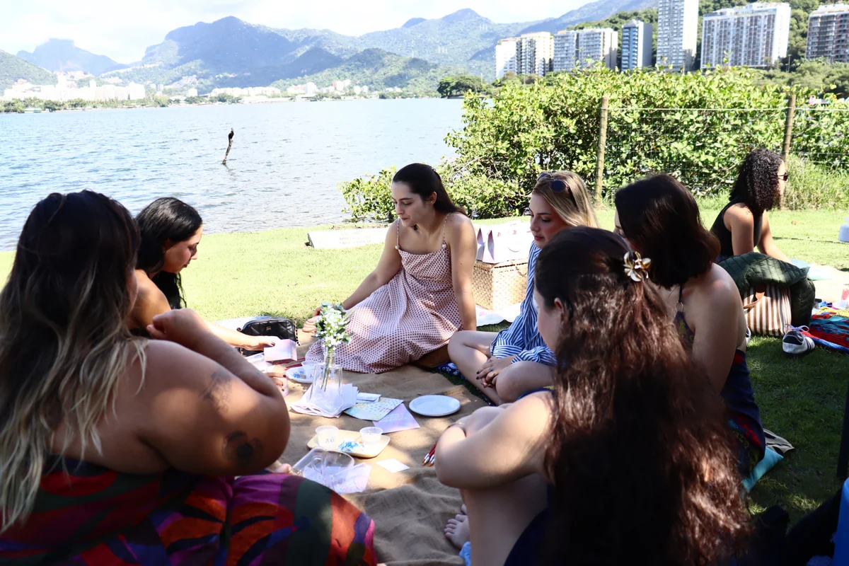 Mulheres reunidas em roda sobre uma toalha de piquenique à beira da lagoa, participando de uma dinâmica de conversa ao ar livre.